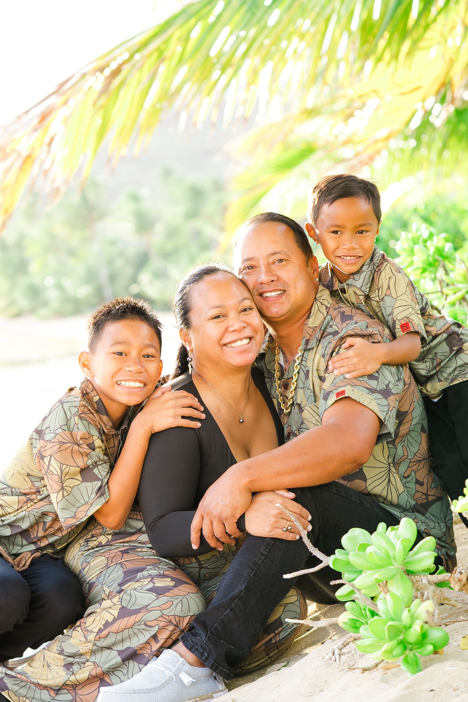 A family wearing matching camouflage outfits sits together under palm trees on a sunny day. Oahu Family Photographer