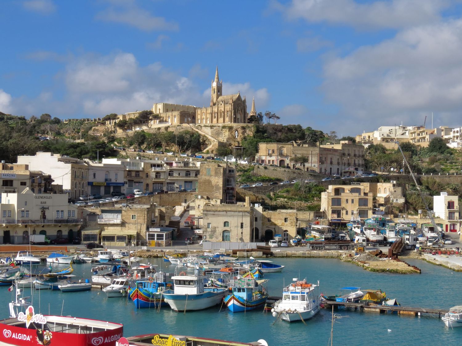 Mediterranean harbor town of Gozo with boats docked in marina and church perched atop hill.