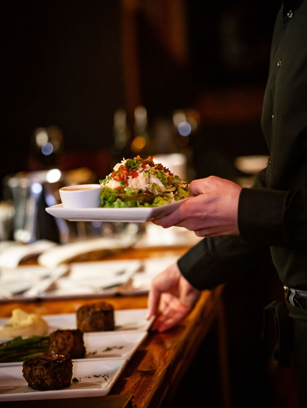 Server hands carrying multiple plates of elegant dinner dishes in dim lighting.