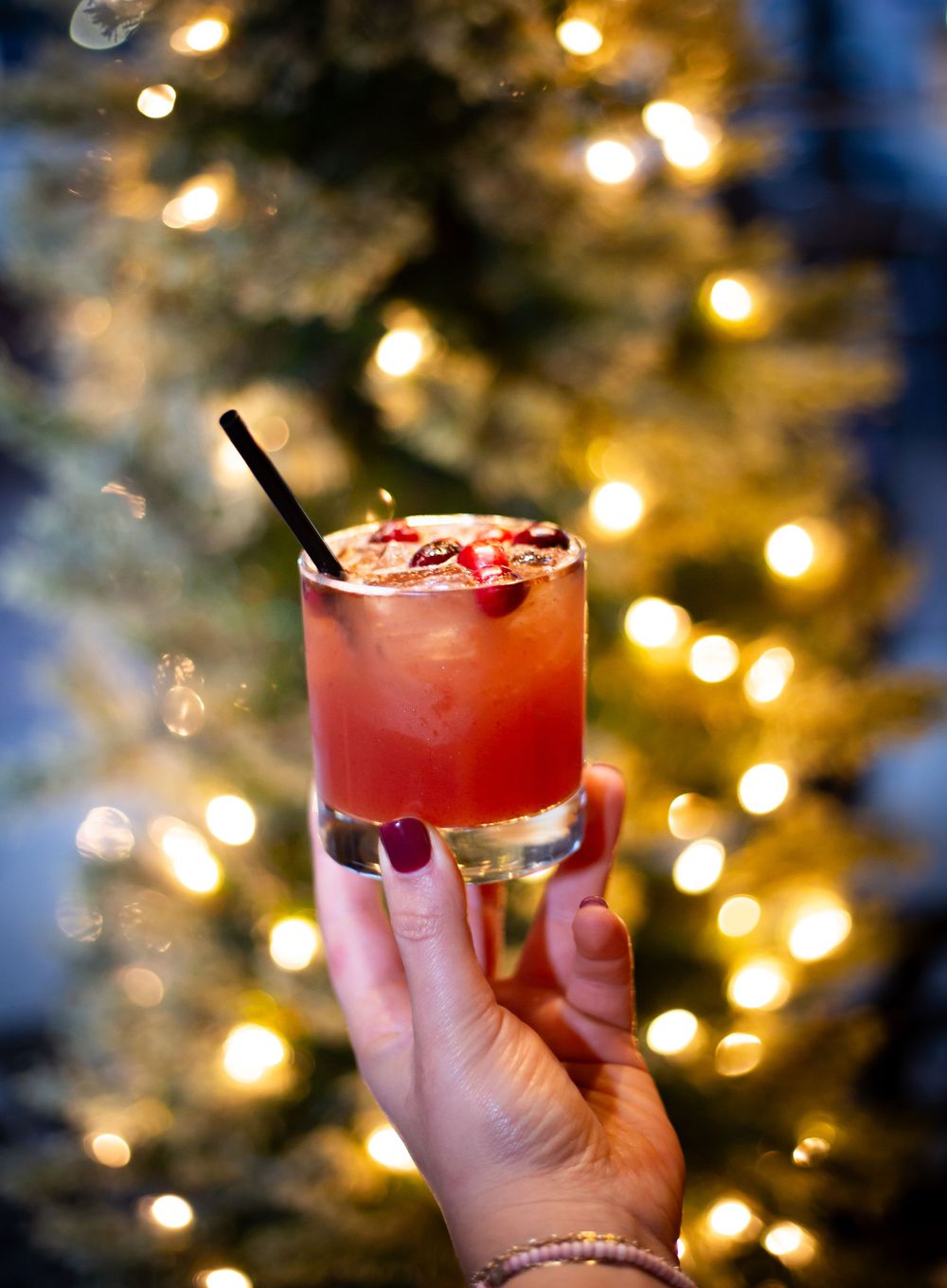 Red cocktail with garnish held against blurred holiday lights background.