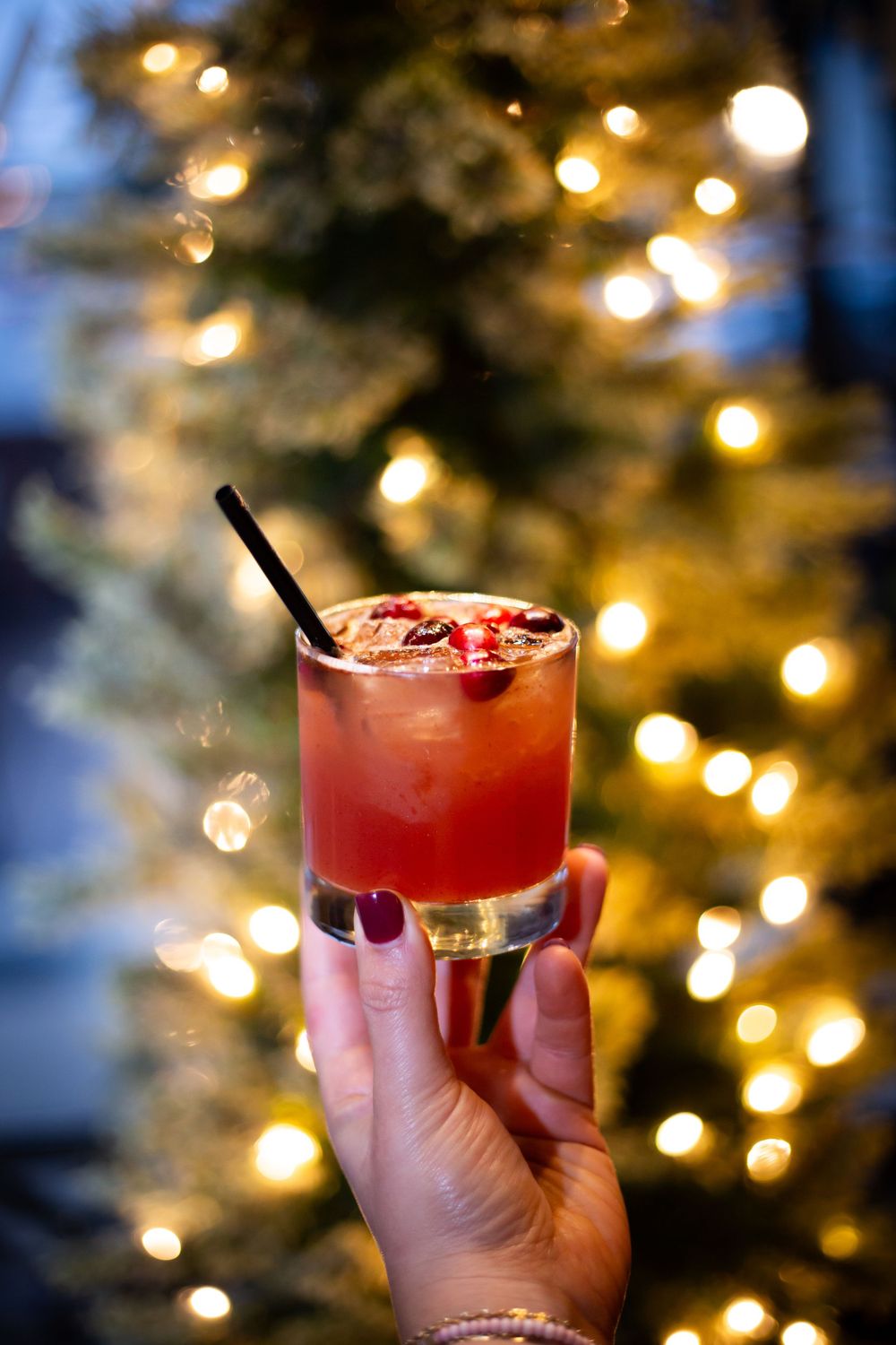 Hand holding a festive red cocktail against bokeh Christmas tree lights.