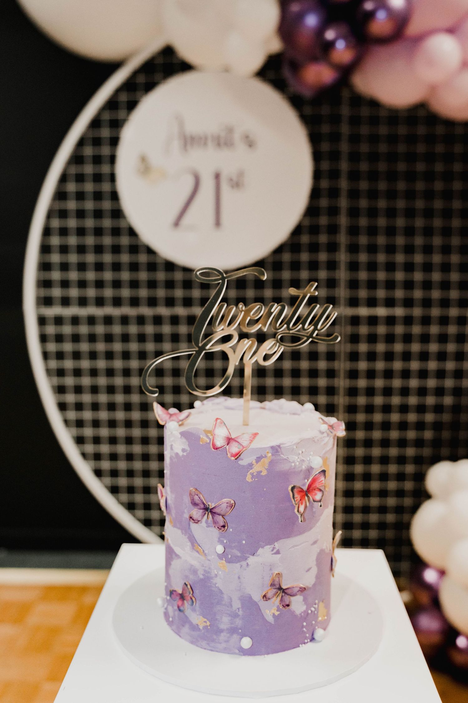 A purple birthday cake decorated with pink butterflies and a gold 'Twenty One' topper on a white circular stand.