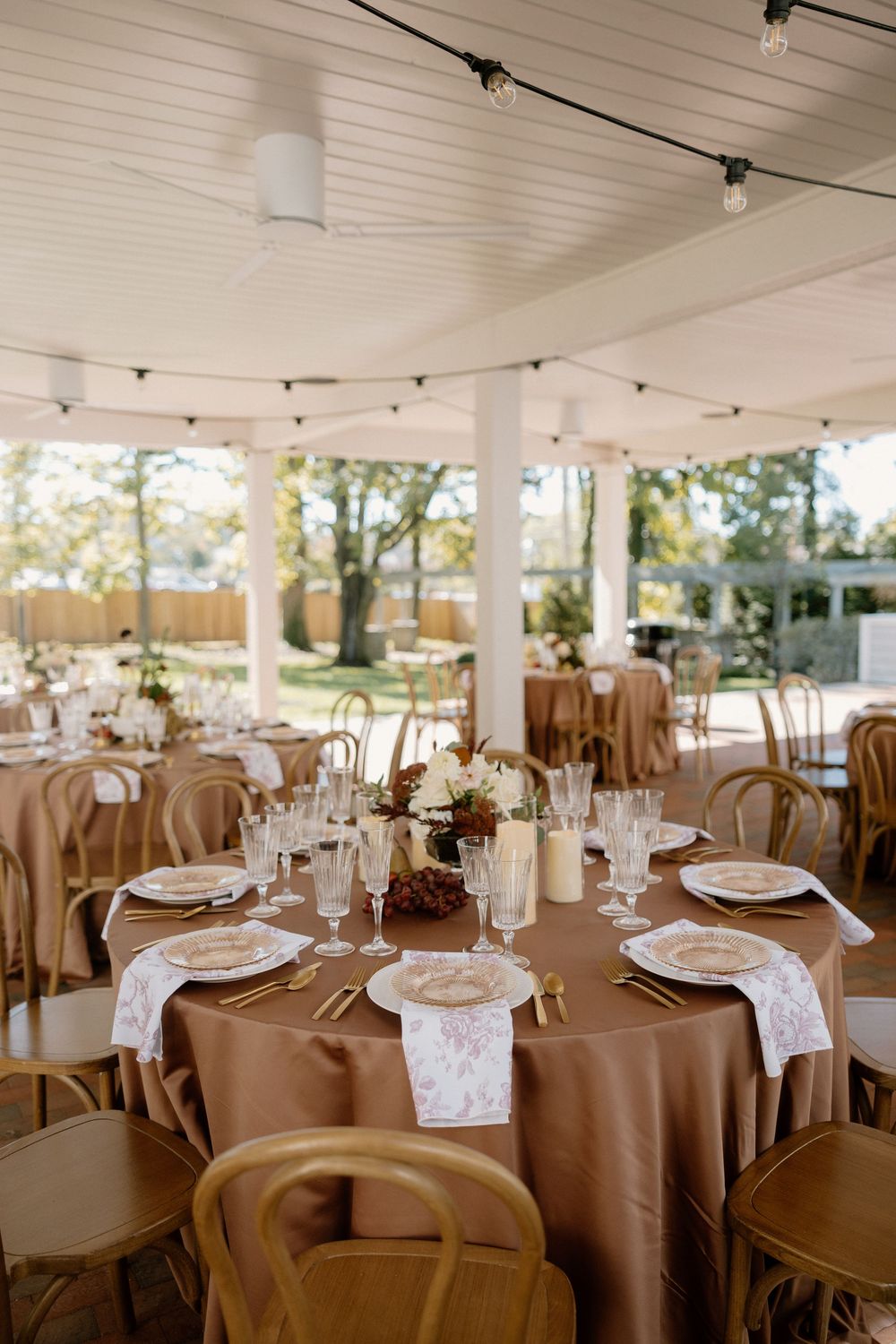 Wedding reception tables set with brown tablecloths and place settings under a covered outdoor pavilion.
