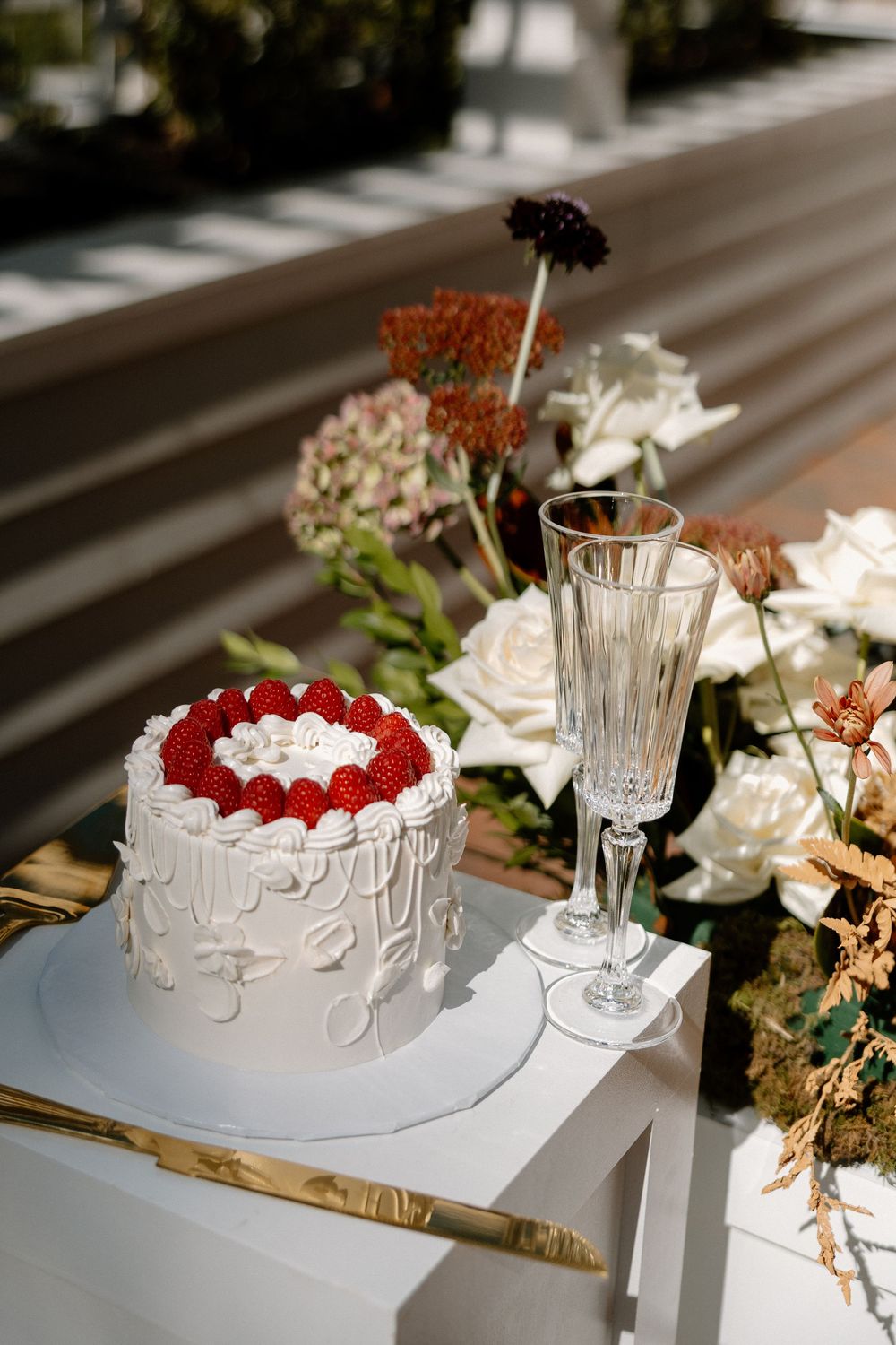 A white frosted cake with red strawberries on top sits on a table with elegant table settings and floral decor.