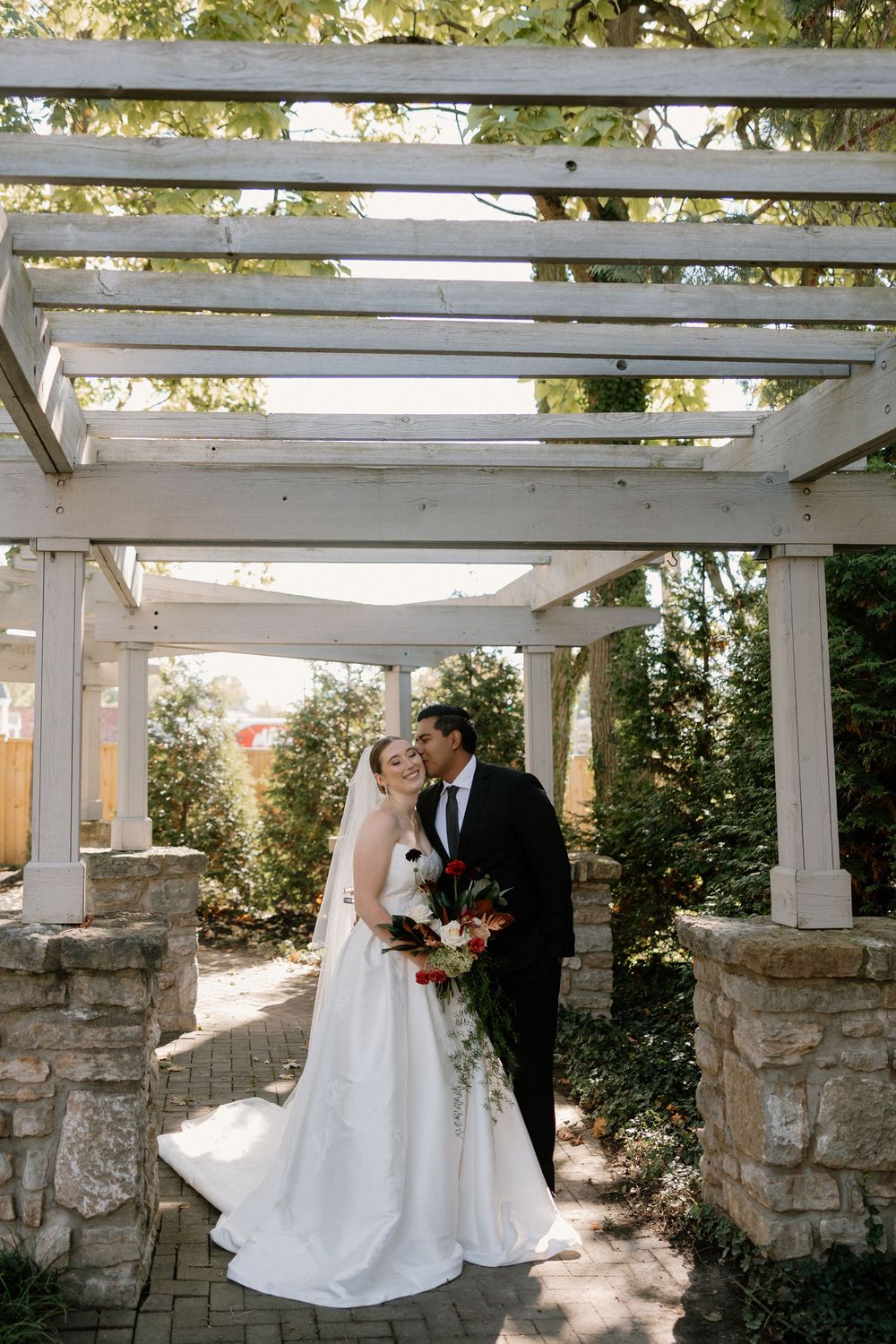 A couple shares a romantic moment under a white pergola in an outdoor garden setting during their wedding celebration.