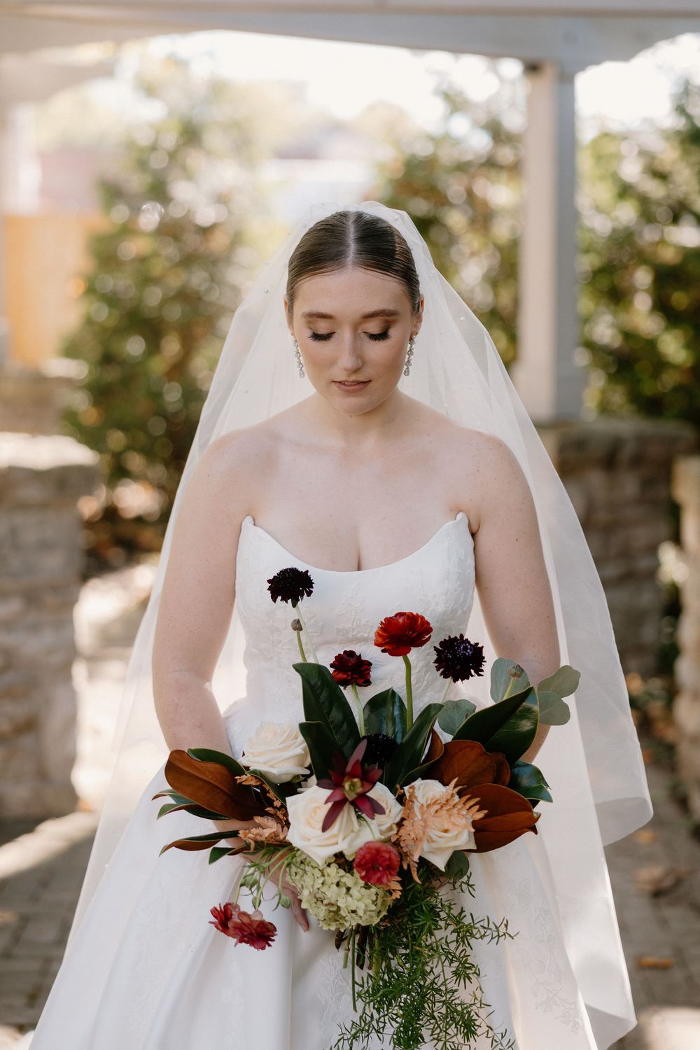 A bridal bouquet features deep red and burgundy flowers with magnolia leaves against a white wedding dress.