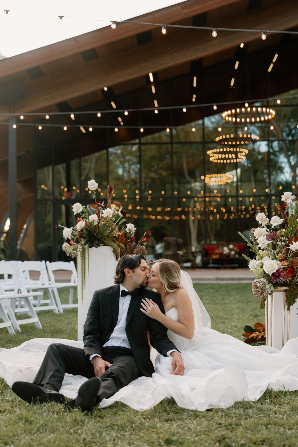 A romantic wedding couple sits together on a lawn at dusk with string lights and floral decorations in the background.