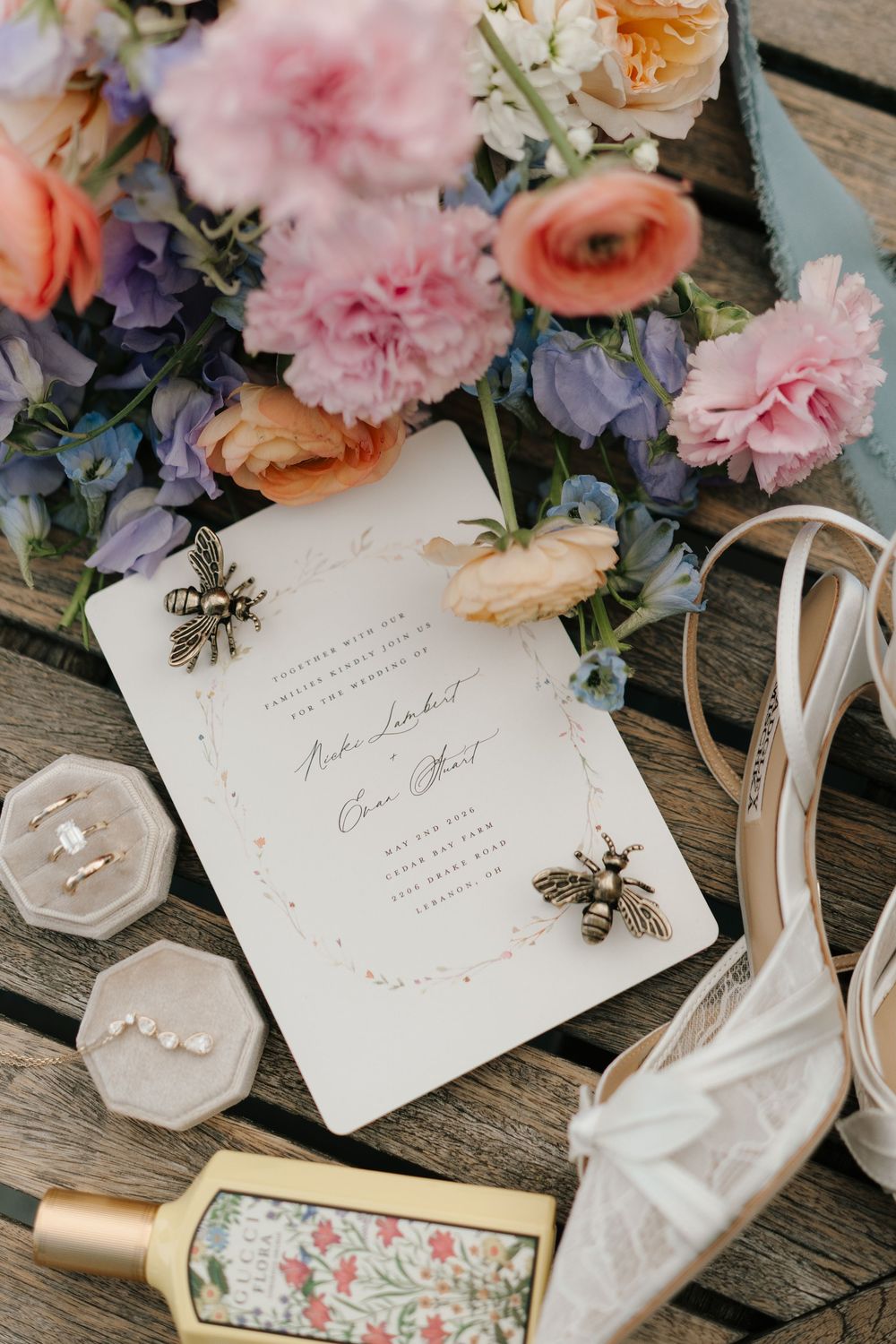 Elegant wedding flat lay with floral bouquet, bee-adorned invitation, ring box, pearl earrings, perfume, and lace bridal heel.