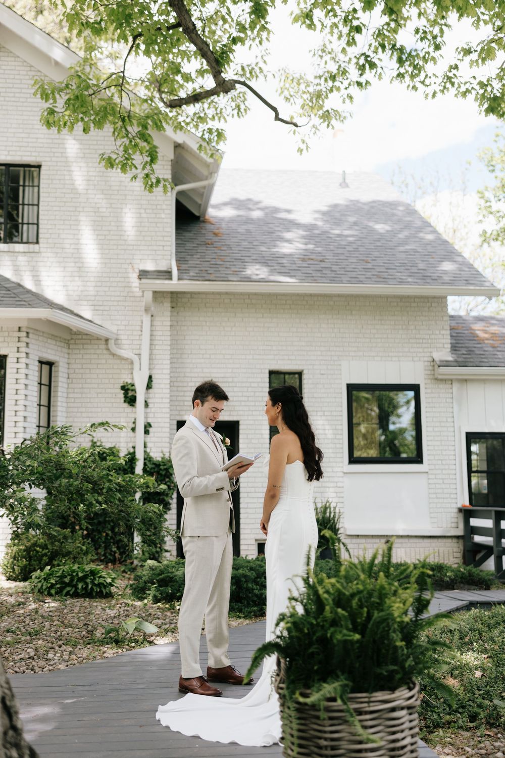 Bride and groom exchange vows outdoors in front of a white brick farmhouse surrounded by lush greenery.