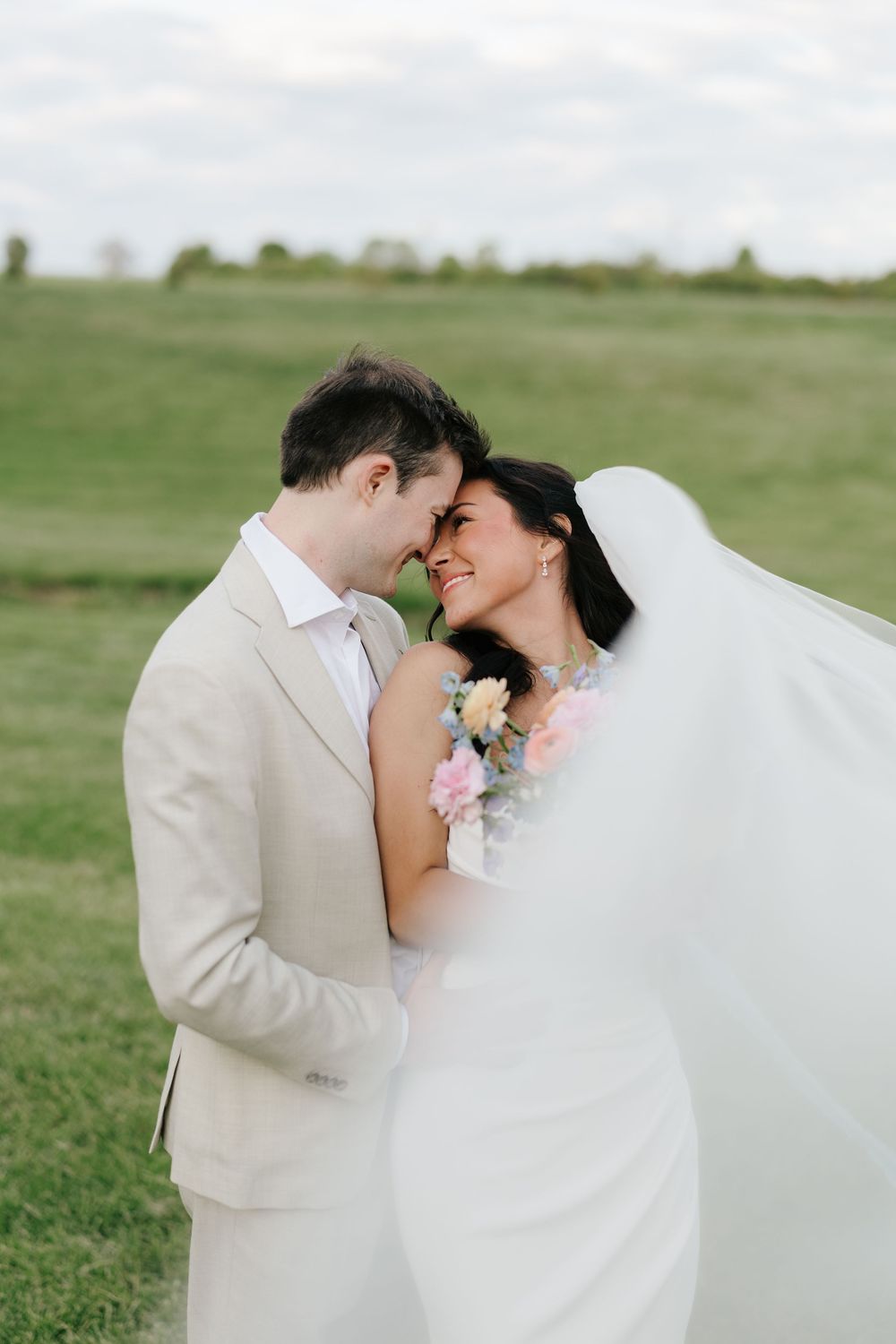 A bride and groom share a romantic forehead-to-forehead moment outdoors, with her veil flowing in a lush green field.