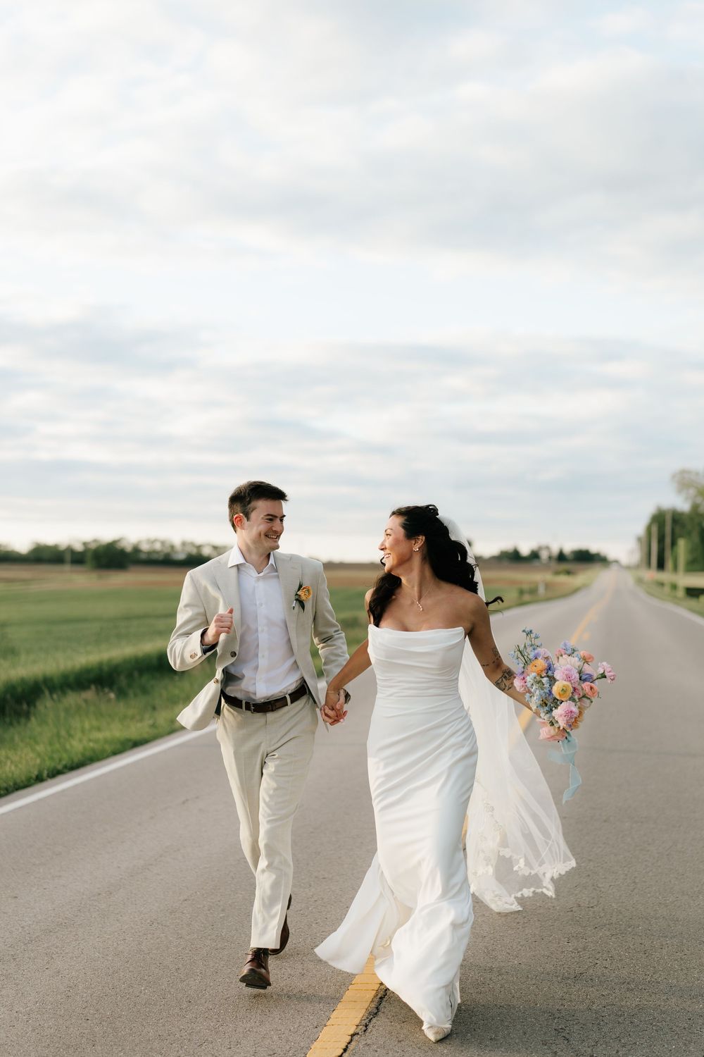 A joyful bride and groom walk hand-in-hand down a rural road, she in a white gown holding a colorful bouquet.