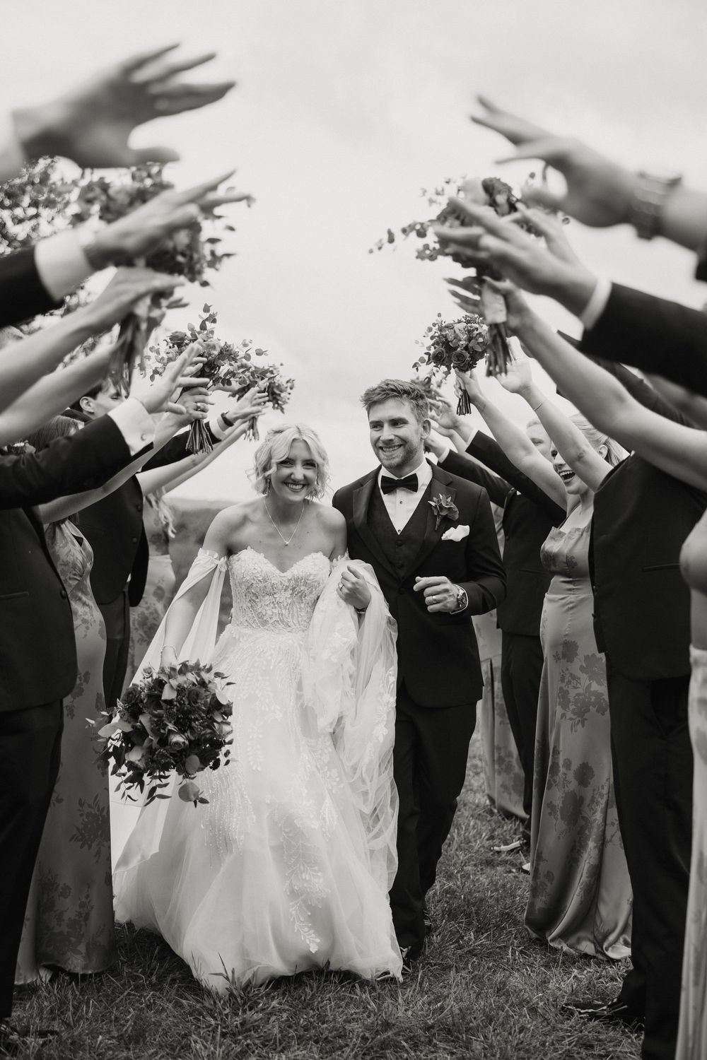 Wedding party forms tunnel with raised arms as newlyweds walk underneath during outdoor celebration.