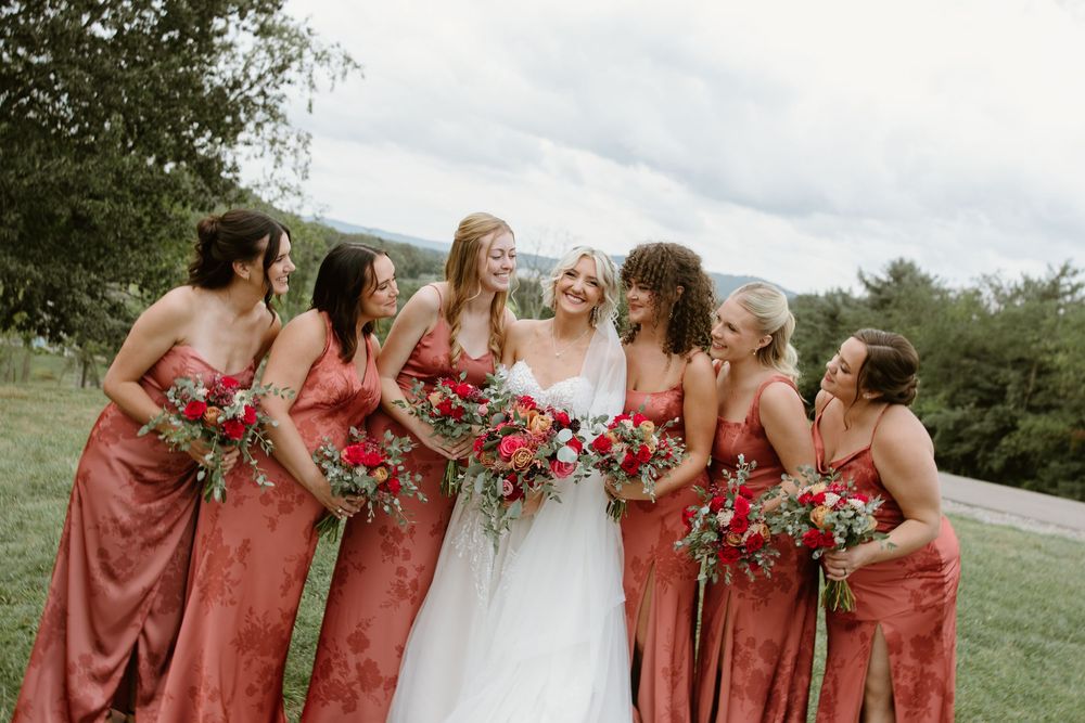 A bridal party in coral-colored dresses holding vibrant red and pink bouquets poses together on a grassy hillside.