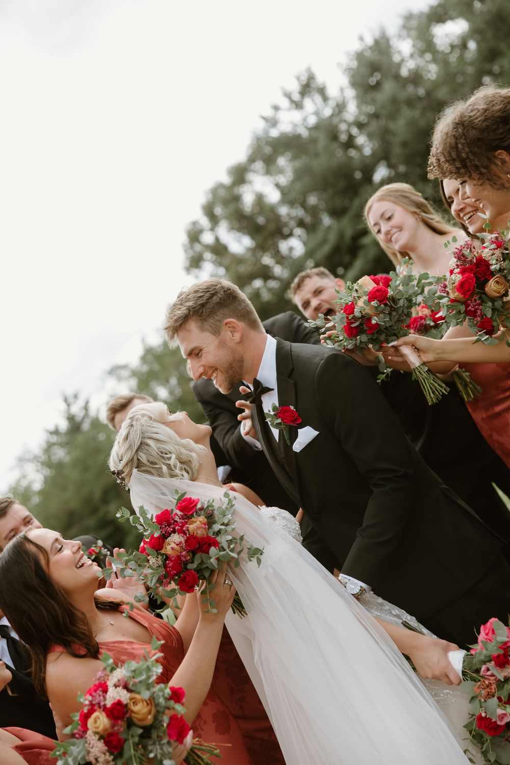 Wedding party poses playfully in a row with vibrant red floral bouquets against a natural backdrop.