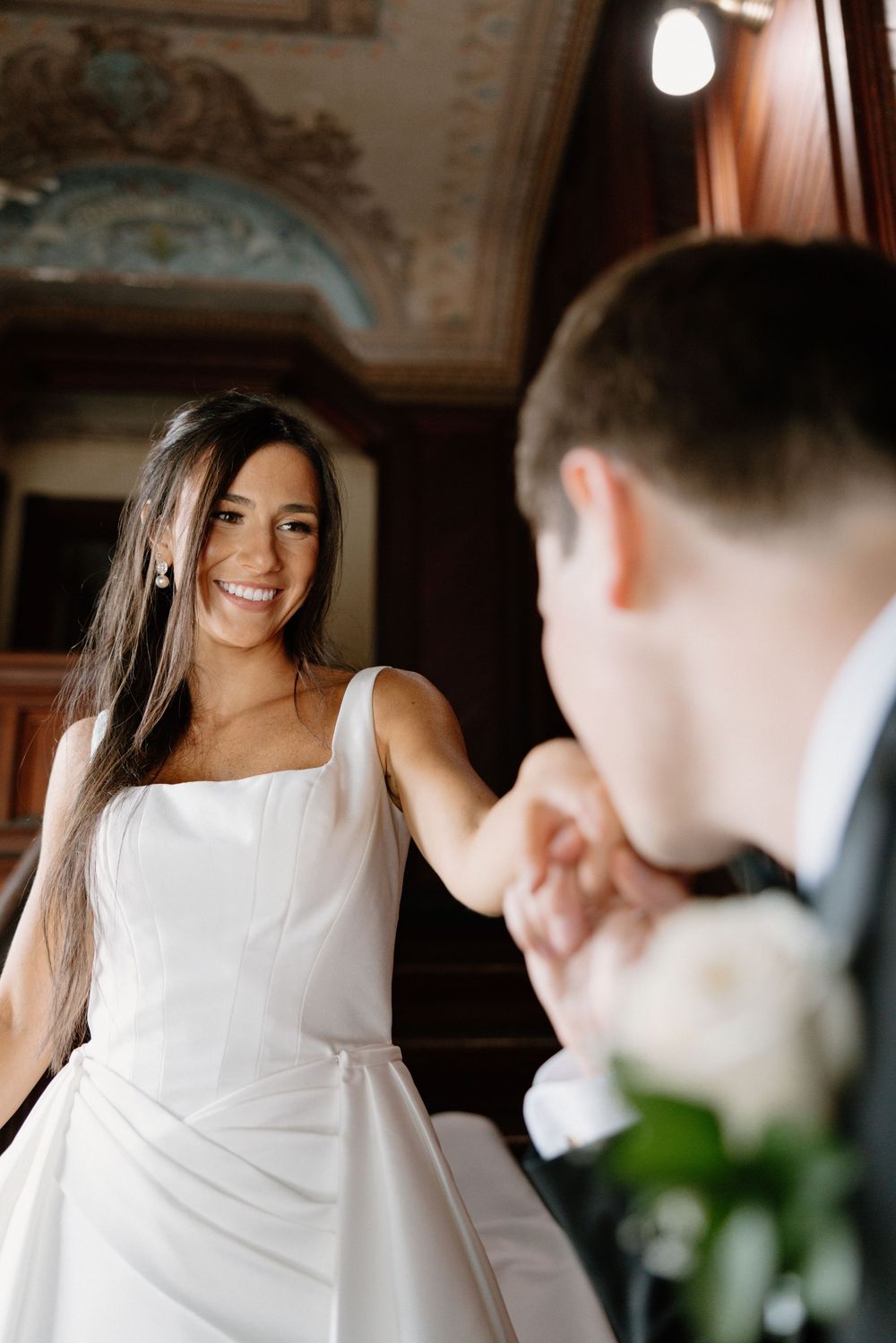 Smiling bride in white gown holds groom's hand in elegant wood-paneled room with ornate ceiling.