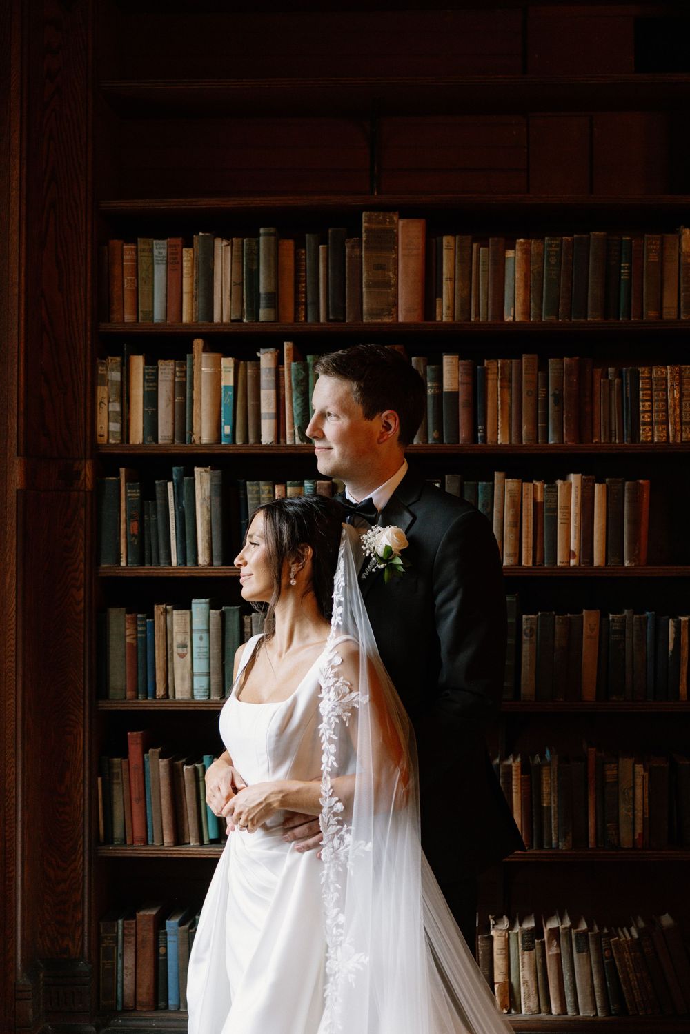 Bride and groom embrace in front of vintage library bookshelves, bride wearing lace veil and white gown.