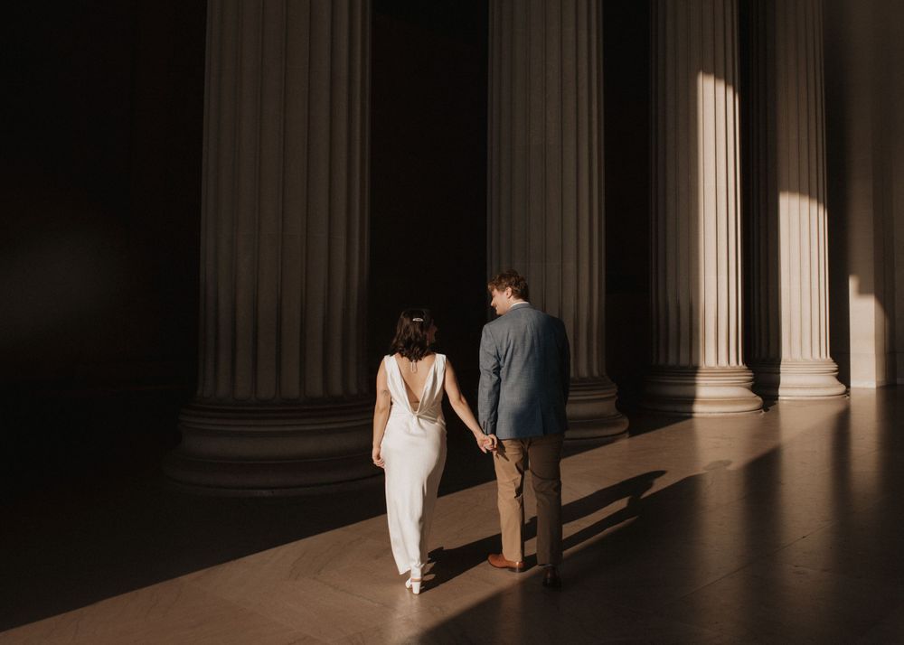 Couple walking through classical pillars with dramatic sunlight casting shadows.