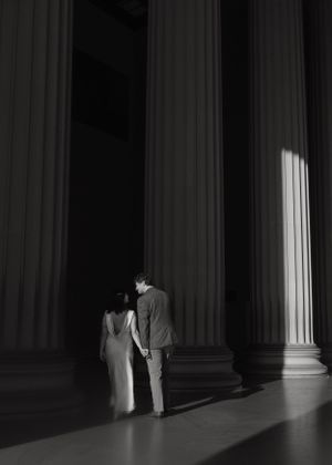 Artistic black and white photo of couple among towering pillars with dramatic lighting.