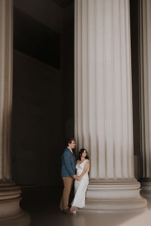 Couple posing intimately against a grand classical column in soft lighting.
