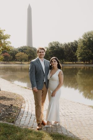 Couple poses on brick path with Washington Monument and reflecting pool in background.