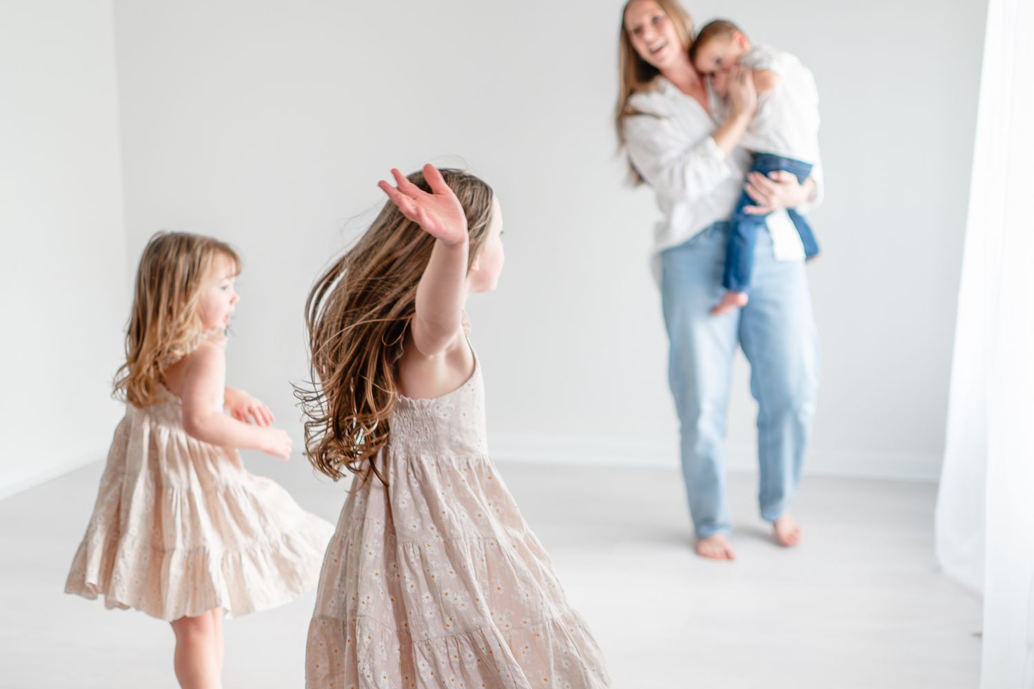 Two children in flowy dresses dance together while a parent and toddler watch from behind in a bright white room.