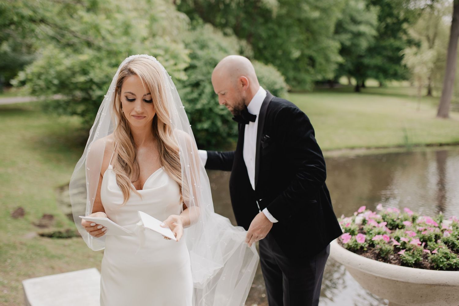 A couple in elegant wedding attire pose together by a pond with pink flowers in the foreground.