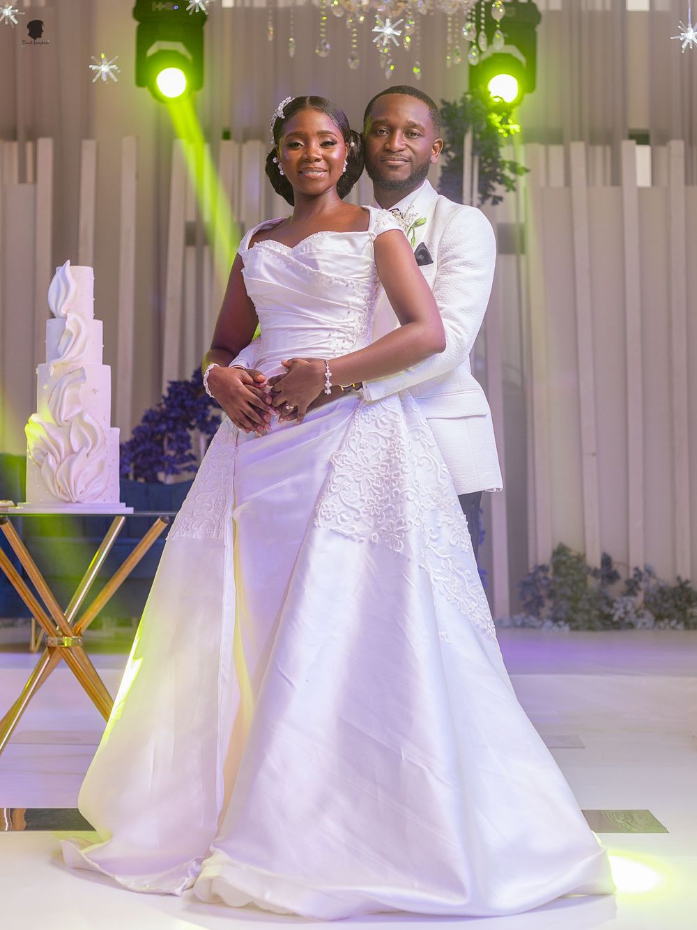 A wedding couple poses together in elegant white attire with a luxurious cake display and green spotlights in the background.