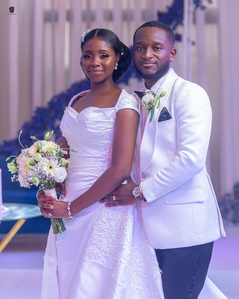 A bride in a white gown holds a pastel bouquet while posing with her groom in a white tuxedo jacket.