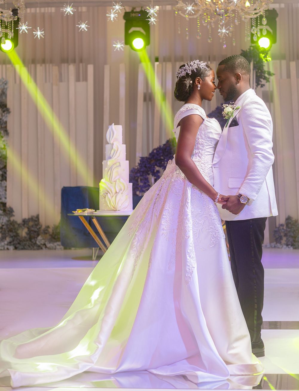 A wedding couple shares an intimate moment by their elegant white cake under decorative green lighting.