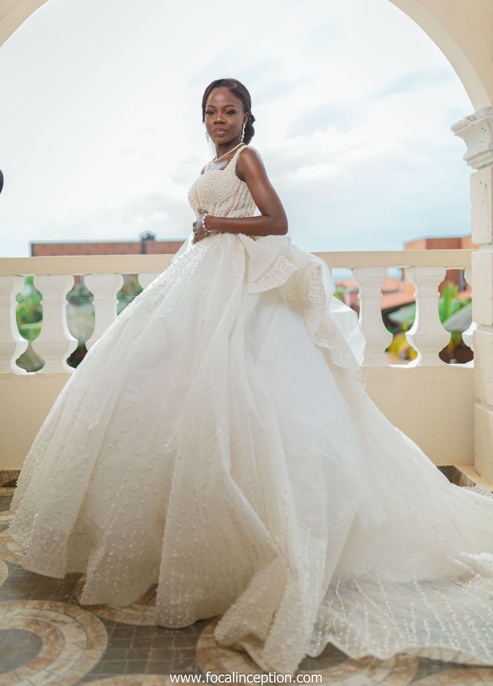 A bride poses on a balcony in a voluminous princess ball gown with intricate details.