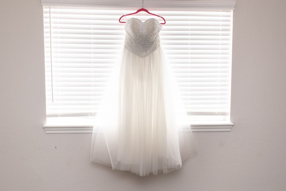 Ethereal white wedding gown with beaded bodice hangs beautifully in front of sunlit window with venetian blinds.