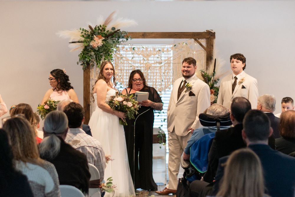 Indoor wedding ceremony taking place under a wooden arch with greenery and floral decorations.