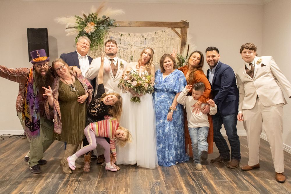 A wedding party celebrating with fun poses in front of a light backdrop in an indoor venue space.