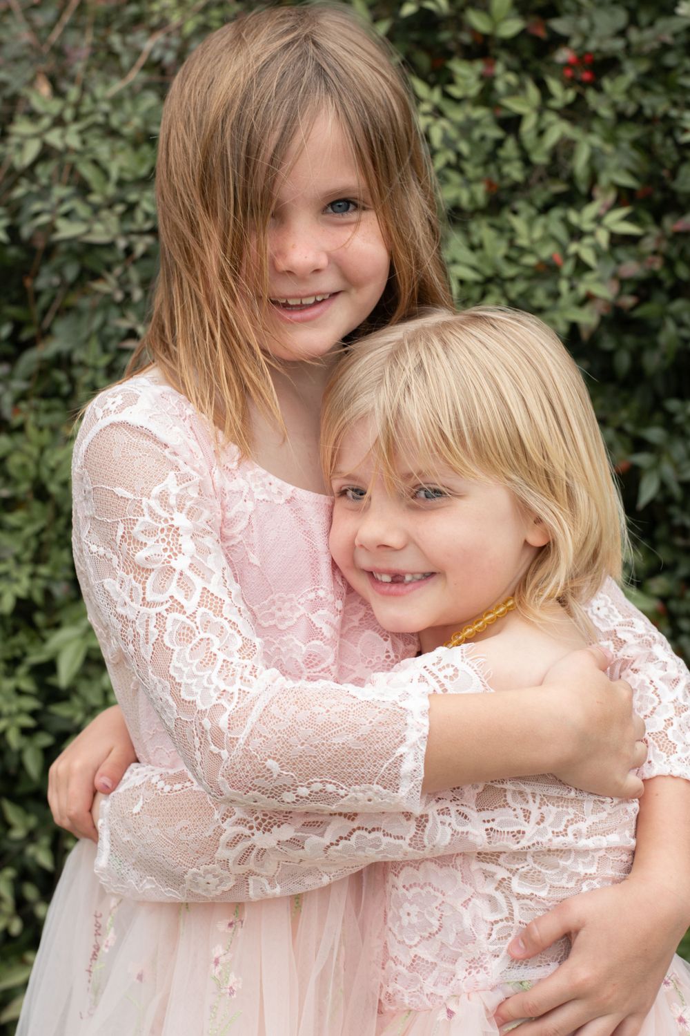 Two children wearing delicate pink lace dresses embrace and smile in front of green foliage.