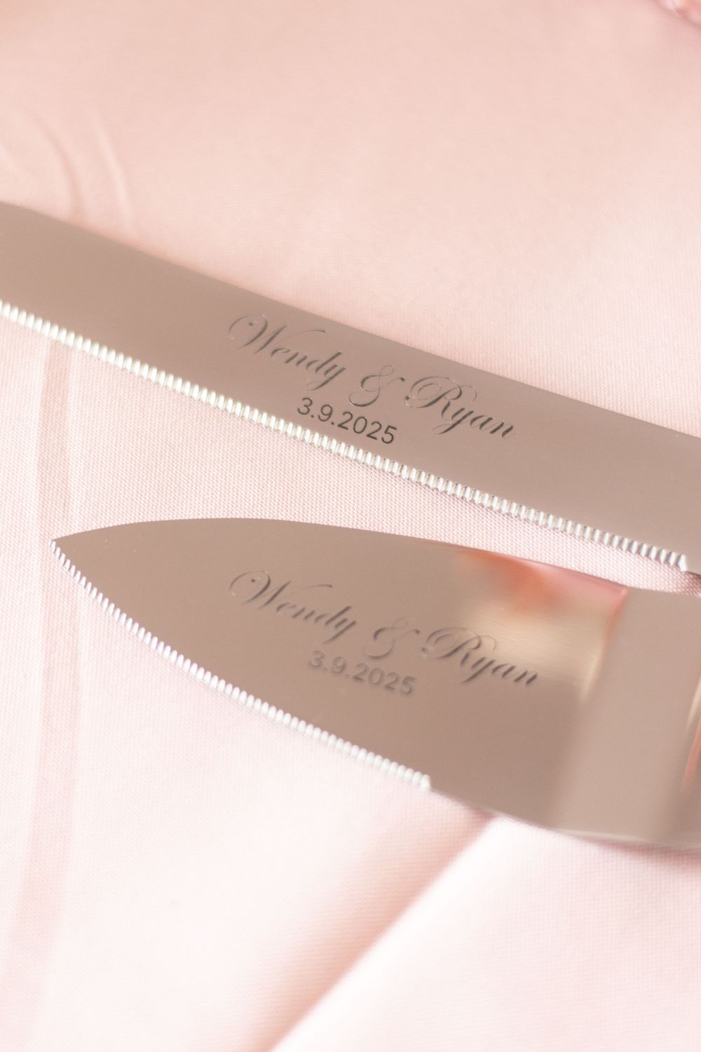 Custom engraved wedding knife set with names and date in elegant script on silver blades against a pink background.