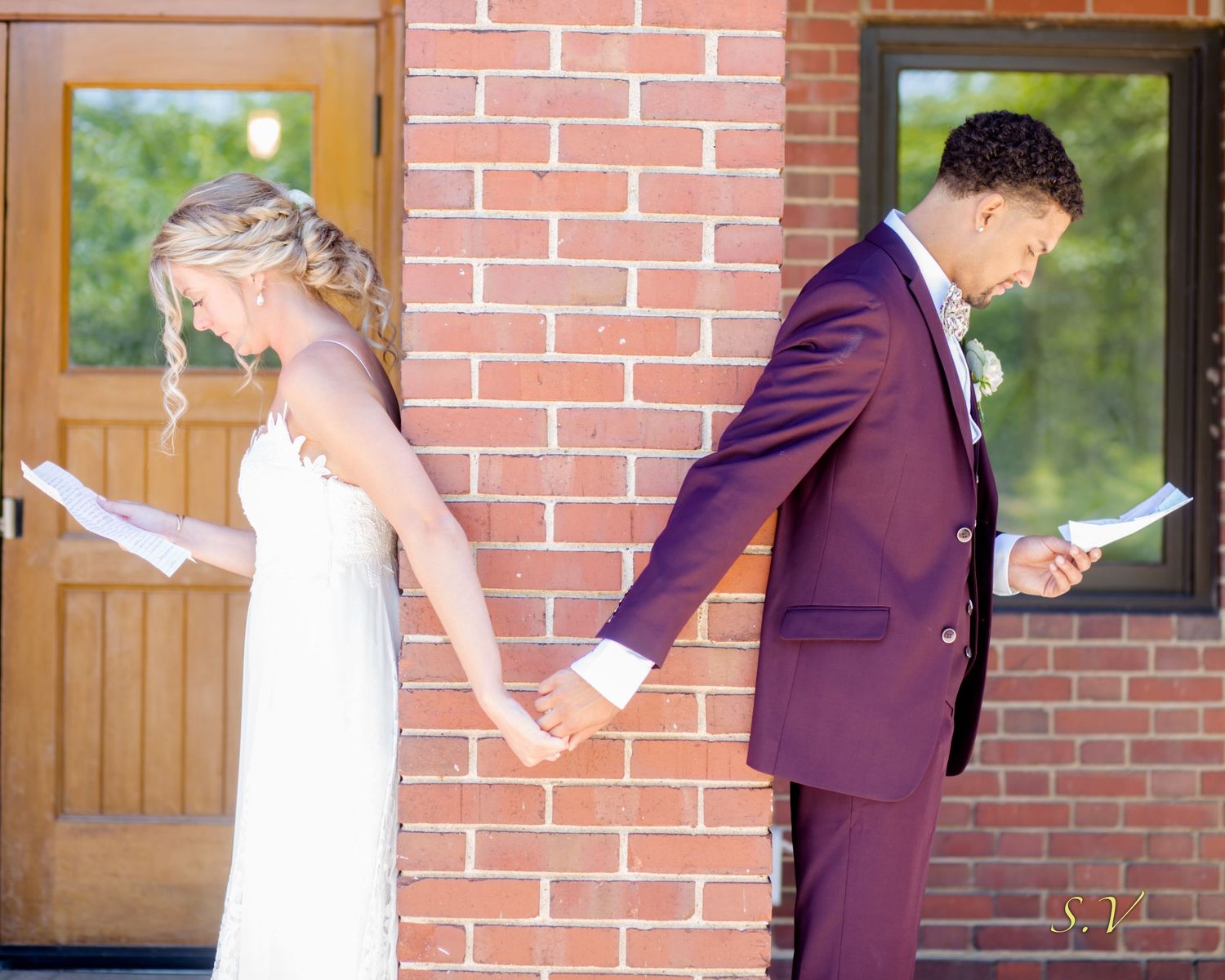 A couple in wedding attire holds hands around a brick corner without seeing each other before their ceremony.