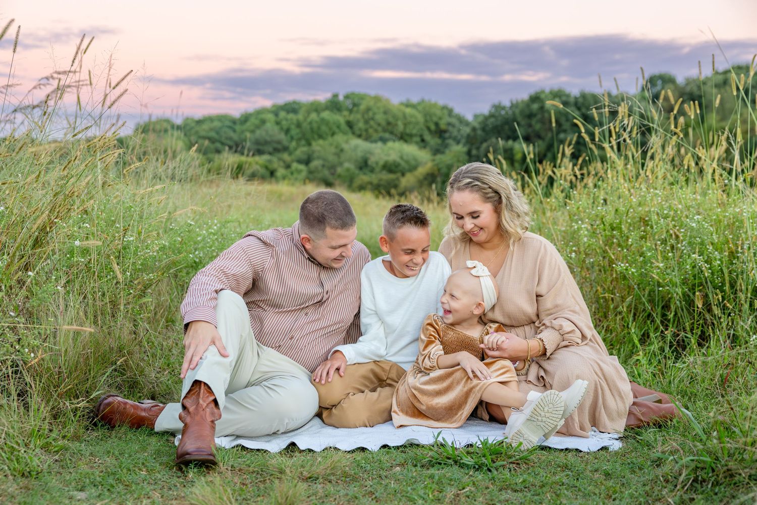 A family in light beige clothing sits together on a blanket in a grassy field at sunset.