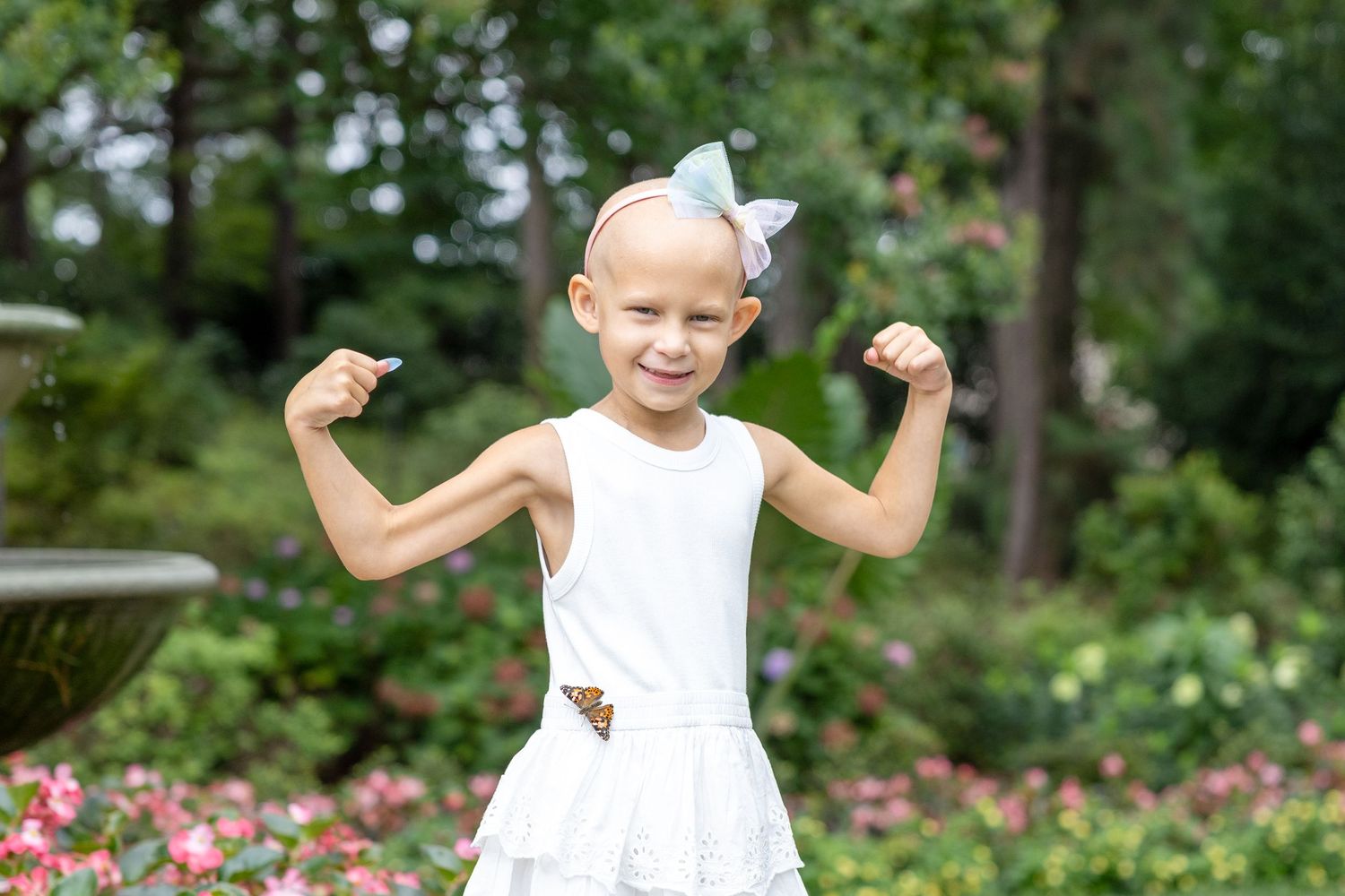 A young child in a white sundress poses with flexed arms in a garden with blooming pink flowers with a Clearwater Butterfly on her dress.