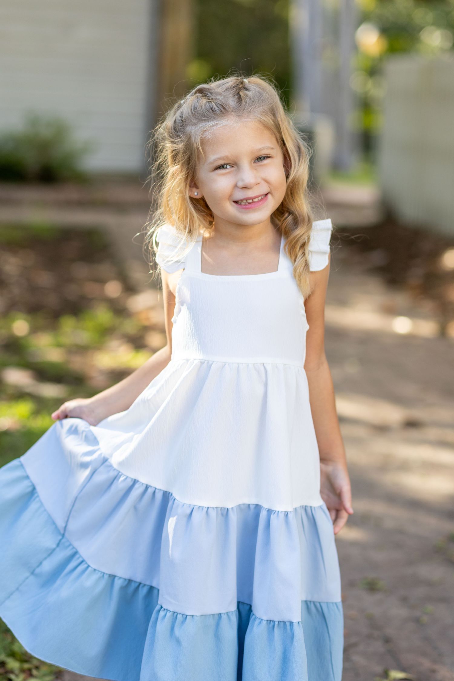 A tiered white and light blue summer dress with ruffled straps features a gradient ombre design in a sunny garden setting.