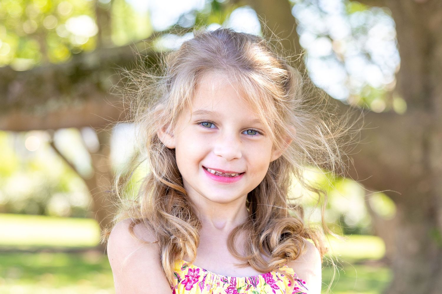 A young child in a floral dress smiles outdoors on a sunny day with wind-blown hair and natural lighting.