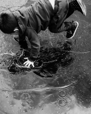 A child in a dark jacket leans forward to touch a frozen puddle with ripple patterns visible beneath the ice.