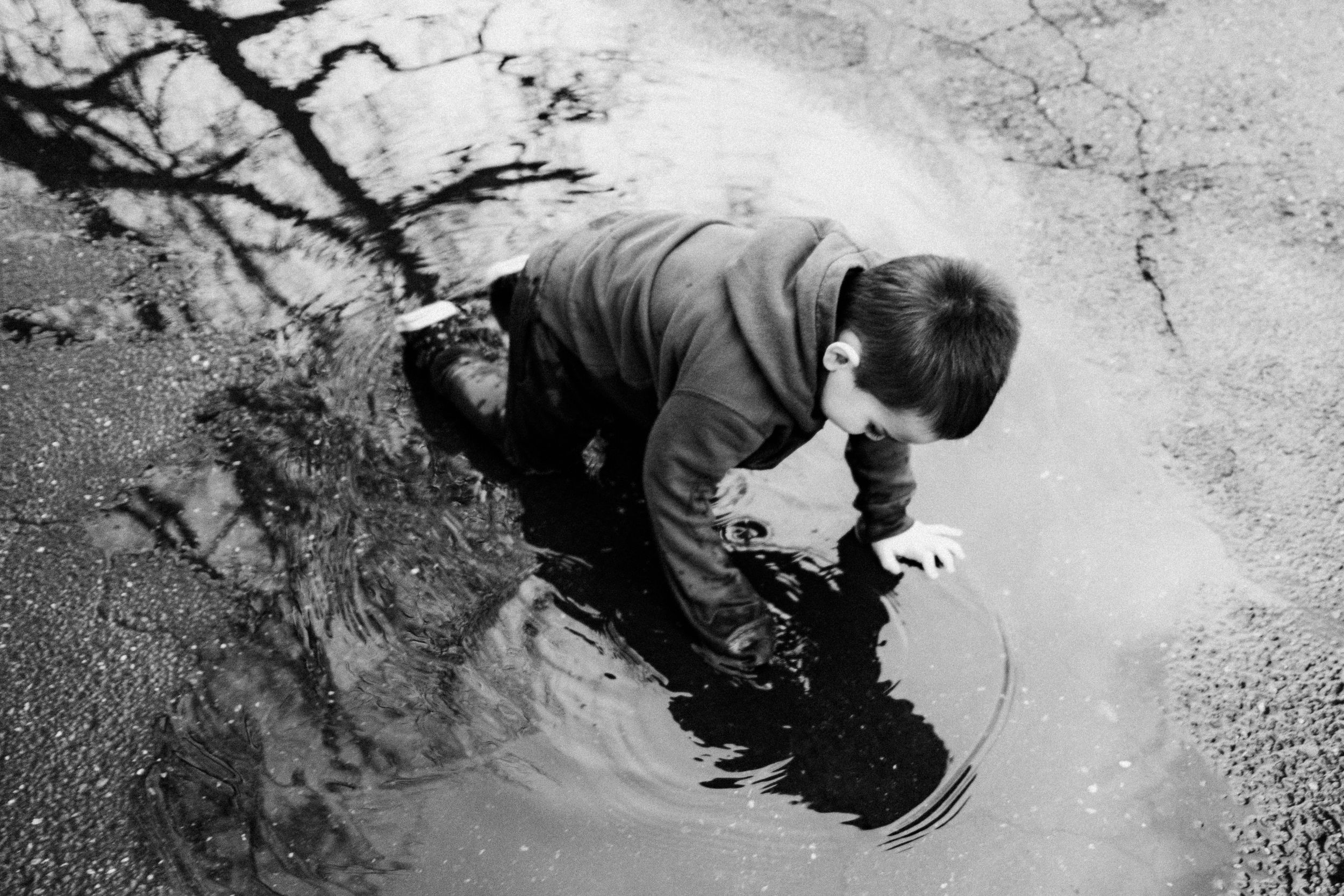 A black and white photograph shows someone bending down to look at their reflection in a puddle of water.
