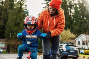 A child learning to ride a bicycle with assistance in orange and blue on a residential street.