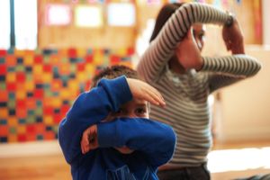 Two young children playing indoors, one in blue covering face while other in stripes stretches arms overhead.