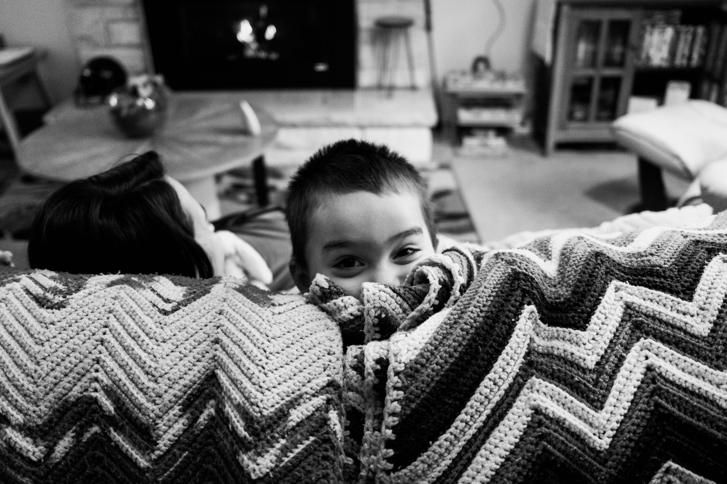 Black and white photo of someone peeking over a chevron patterned blanket in a cozy living room setting.