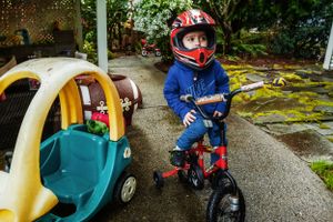 A small child riding a red tricycle while wearing a red helmet and blue outfit next to a toy car.