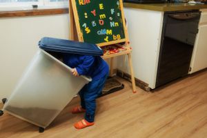Small child bending over and looking behind furniture in a classroom setting with colorful educational board visible.