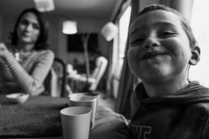 A black and white photo shows a small child smiling next to a cup on a table while someone sits in the background.