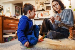 A child in a blue sweater and adult in stripes sit together on the floor of a cozy home interior.