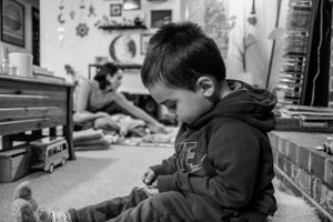 A young child in a hooded jacket concentrates intently while playing in a cluttered indoor setting in black and white.